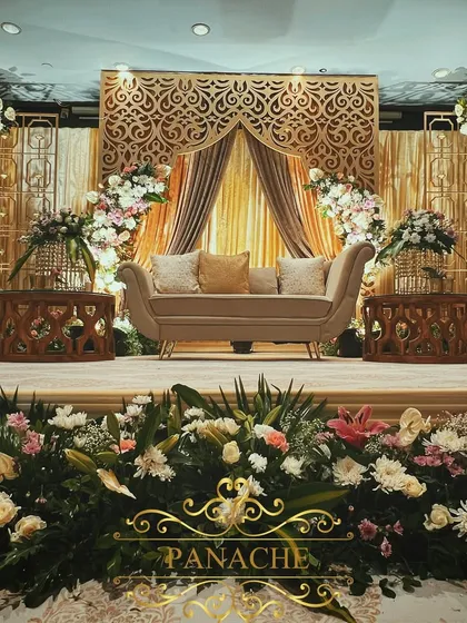 The main stage for the Roka, featuring a backdrop of intricate gold laser-cut panels and soft, flowing drapes. The stage is framed with large floral arches, creating a picture-perfect setting.