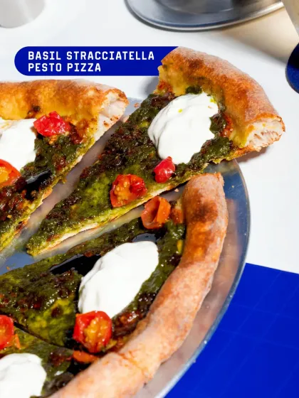 A slice of our Basil Stracciatella Pesto Pizza, featuring a vibrant green pesto base, roasted cherry tomatoes, and creamy stracciatella.