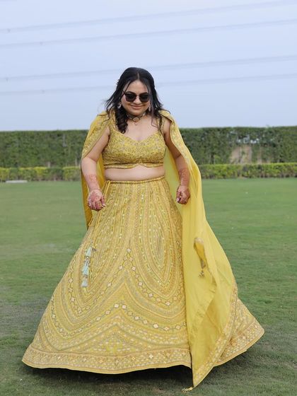 The bride is bringing the main character energy to her mehendi. The cape-style dupatta and sunglasses add a modern, playful touch to her embroidered lehenga.