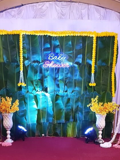 A traditional South Indian style baby shower backdrop made from fresh banana leaves. The look is completed with marigold flower garlands and a 'Baby Shower' neon sign.
