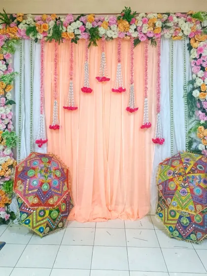 A beautiful backdrop for a home function, featuring peach drapes, floral hangings, and colorful Rajasthani umbrellas.