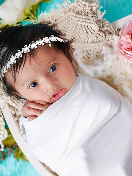 A beautiful portrait of an awake newborn with striking eyes, swaddled in white and surrounded by soft flowers against a bright aqua background.