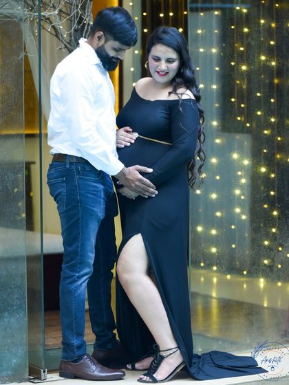 An elegant evening shot of a couple by a glass wall with fairy lights. The mom-to-be looks stunning in a black off-shoulder gown.