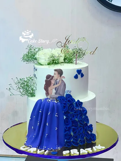 A grand two-tier engagement cake with a flowing blue gown design. The cake features an edible print of the couple, with the bride's dress cascading down the side in beautiful blue ruffles.