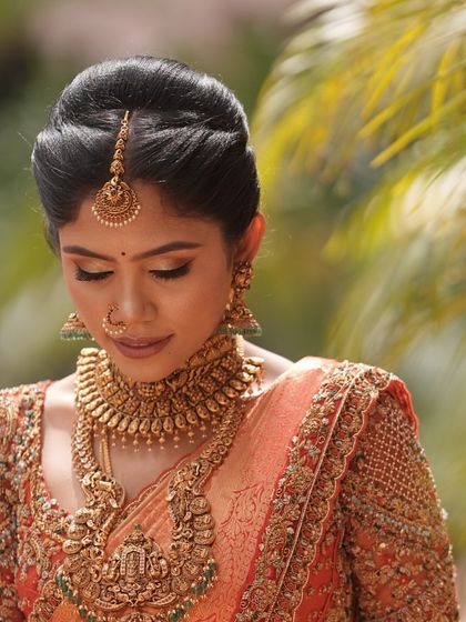 A portrait of a bride in a traditional orange Kanjivaram saree. The heavily embroidered blouse and antique gold temple jewelry create a look of divine elegance for her wedding day.