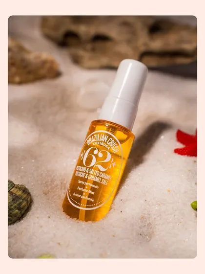 A close-up of a perfume mist nestled in the sand. The warm, golden light and soft focus create a dreamy, sun-kissed aesthetic for this summer fragrance product.