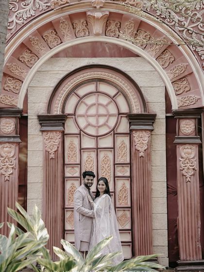 A wide shot showcasing the couple in their traditional outfits against a backdrop of a massive, beautifully designed arched doorway.