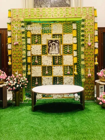 A beautiful pooja or engagement setup with our signature checkerboard backdrop. The green and gold jaali frame adds a touch of luxury, while the floral arrangements bring freshness.