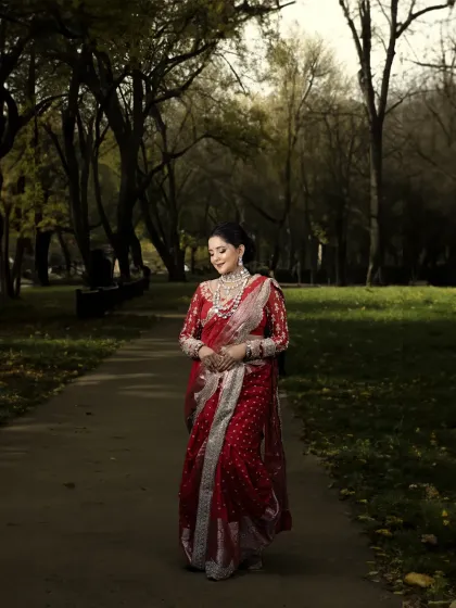 A full-length portrait of a bride in a striking red saree, walking through a park. The moody lighting and natural setting create a cinematic feel.