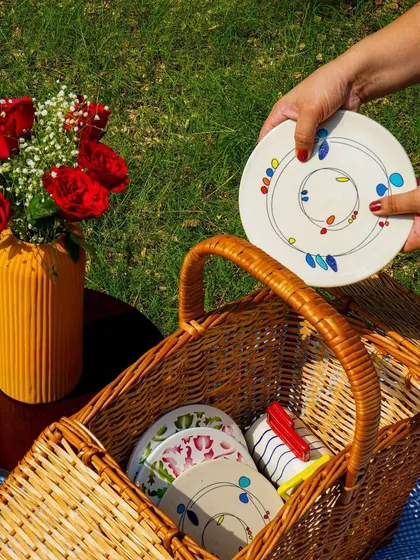 A lifestyle shot of a picnic scene, with a hand placing a plate into a wicker basket. This tells a story and shows the products in use.