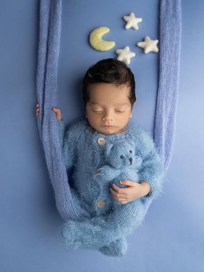 Floating on a cloud. This baby boy, dressed in a fuzzy blue outfit, sleeps in a fabric swing prop against a solid blue background with a moon and stars.