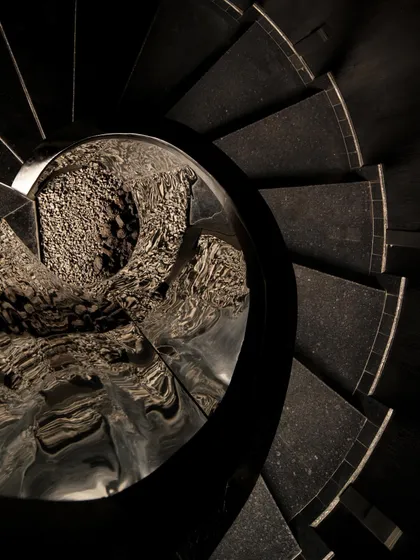 An abstract, top-down view of the staircase, where the steps create a hypnotic spiral. The reflection of the textured ceiling in the central column adds another layer of complexity to the visual experience.