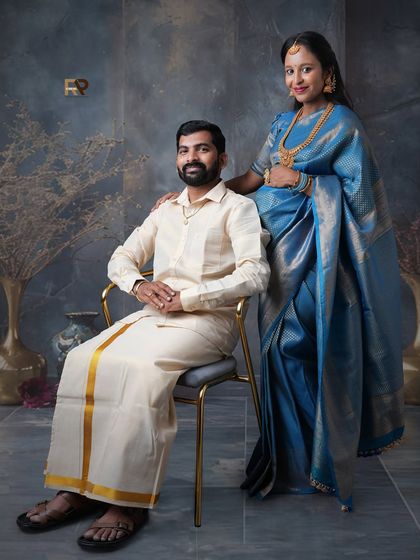A regal portrait of an expecting couple in traditional South Indian attire. The husband is seated, and his wife stands beside him, creating a formal and dignified composition.