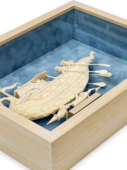 The Sholapith boat is set against a baby blue suede mat, evoking a sense of water. The maple veneer shadow box frame provides a sturdy and elegant enclosure for this delicate piece.