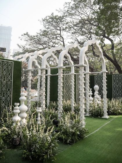 An entryway of white arches and sculptural elements, surrounded by greenery, creates a grand and artistic entrance for the 'Oasis of Love' ceremony.
