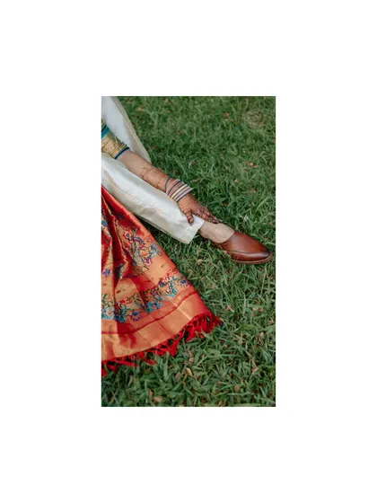 An artistic detail shot focusing on the bride's colorful saree and the groom's traditional jutti shoes, resting on the green grass.
