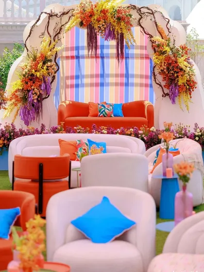 A vibrant and modern lounge area for a wedding event. The mix of orange and white furniture with bright blue cushions and a colorful striped backdrop creates a playful and artistic vibe.