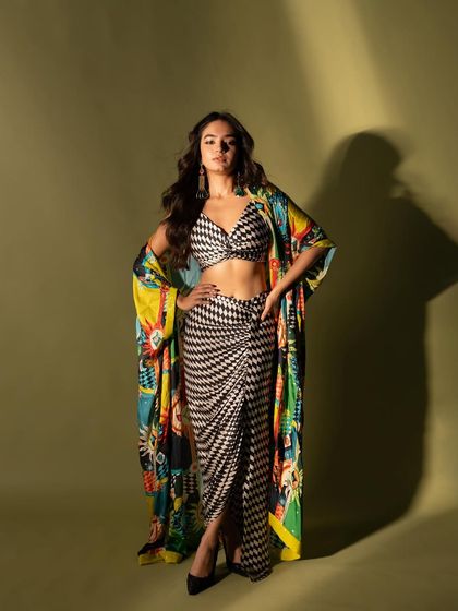 A full-length studio shot of Anushka Sen in a vibrant, patterned outfit. The hard light creates a strong shadow, adding a graphic element to the portrait.