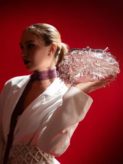 A high-fashion look featuring a model holding a tinsel clutch. The styling, with a white blazer and statement accessories, combined with the dramatic red background, creates a powerful and memorable campaign image.