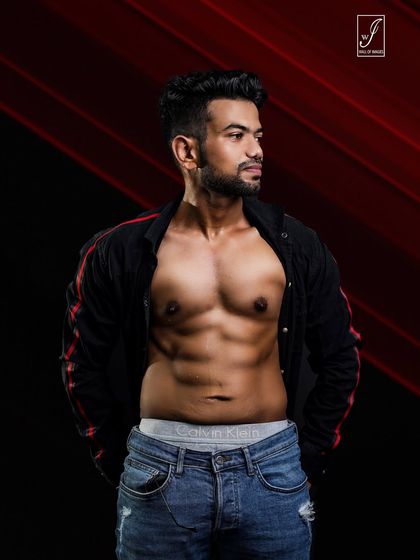 A relaxed yet strong pose with an open shirt, revealing a well-defined chest and abs. The red-accented background adds a touch of color and energy to this studio fitness portrait.