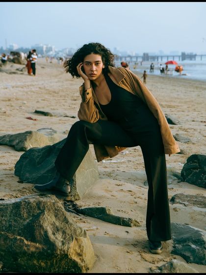 A strong, confident pose on the rocks. The composition balances the model with the expansive beach scene in the background.