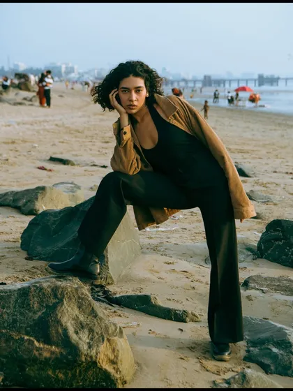 A strong, confident pose on the rocks. The composition balances the model with the expansive beach scene in the background.