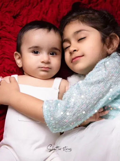 A heartwarming shot of an older sister hugging her baby brother, both lying on a soft red blanket.