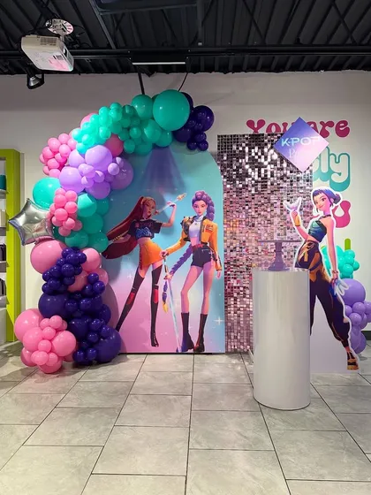 A K-pop themed birthday decoration featuring character standees, a silver sequin panel, and a vibrant balloon arch in shades of purple, pink, and teal.