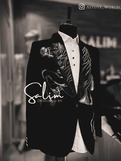 A mannequin displaying a black tuxedo jacket with an abstract, wing-like embroidery design on the lapel. The black and white photo emphasizes the sharp lines of the artwork.