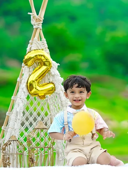 A happy birthday boy holds a yellow balloon, sitting by a macrame teepee during his outdoor second birthday celebration.