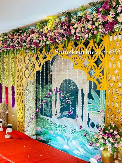 A fusion decor backdrop combining a traditional Pichwai-style painting with modern gold lattice panels. The top is adorned with a lush arrangement of colorful flowers.