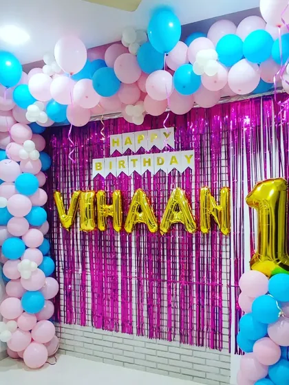 A first birthday decoration using a vibrant mix of pink and blue balloons. The setup includes a full balloon frame around a pink shimmer curtain, with a large gold '1' foil balloon on a pillar.