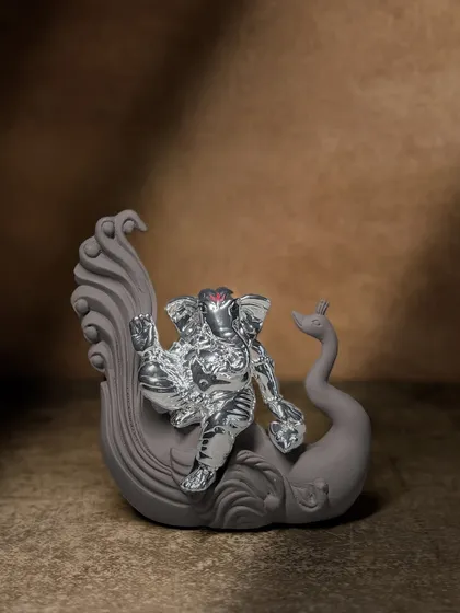 A modern and artistic representation of Lord Ganesha, seated on a gracefully sculpted peacock. The matte grey finish contrasts beautifully with the silver-plated idol, making it a contemporary piece of spiritual art.