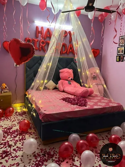 A cozy and romantic birthday room surprise. The setup features a bed with a light-up canopy, "HAPPY BIRTHDAY" foil balloons, a large teddy bear, and the floor covered in red and white balloons with rose petals.