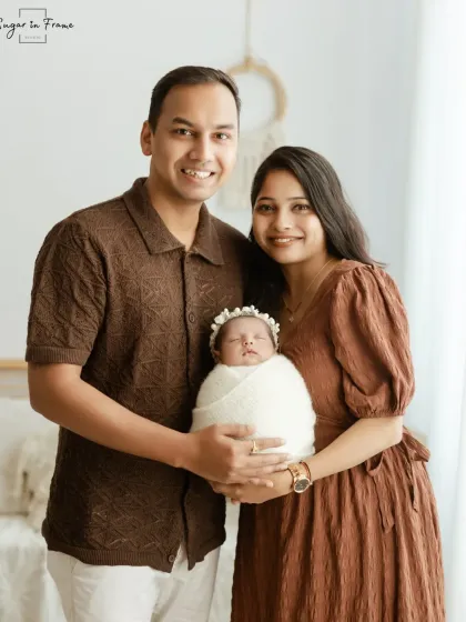 A classic, smiling family portrait. The new parents hold their swaddled baby and smile directly at the camera, a perfect photo for sharing with loved ones.