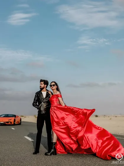 A high-fashion, editorial style shot on a desert road in Dubai. The luxury car, the flowing red dress, and the confident poses come together to create a powerful image straight out of a magazine.