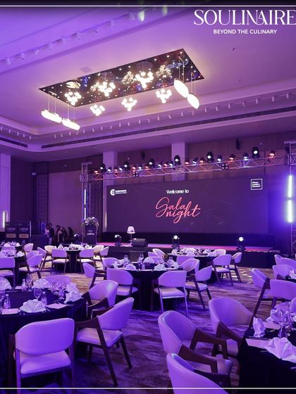 A wide shot of a gala night venue, with tables set under sophisticated purple lighting. We create an ambiance of refined charm for large-scale sit-down dinners and corporate events.