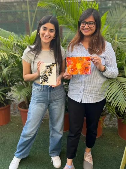 Two friends posing with their artwork against a backdrop of lush green plants. A perfect blend of creativity and nature.