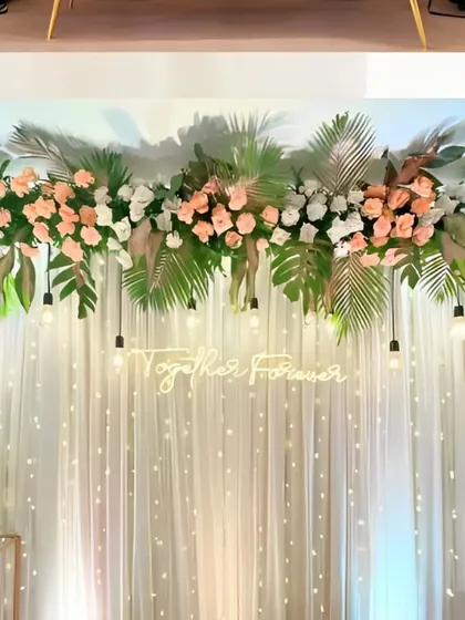 A minimalist and tropical-inspired stage design, featuring a 'Together Forever' neon sign, a canopy of monstera leaves and peach roses, and a curtain of twinkling fairy lights.