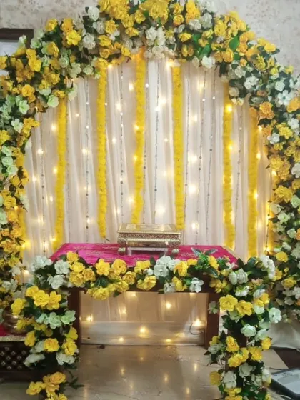 A beautiful floral arch for a home pooja or intimate function. The arch is made of yellow and white flowers, set against a backdrop of white curtains and fairy lights.