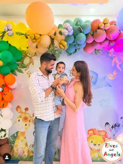 A happy family photo at Vidit's colorful first birthday party. The backdrop features cute woodland creatures and a rainbow balloon arch.