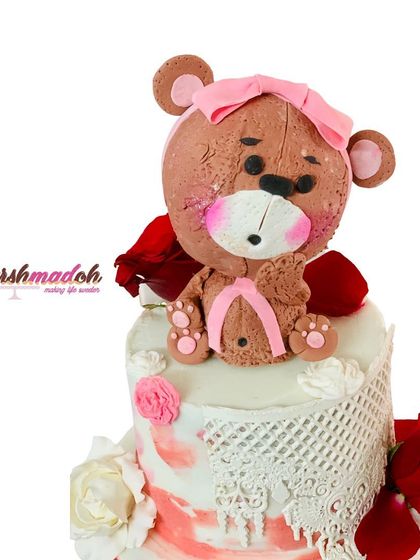 A close-up of the 3-tier first birthday cake, focusing on the intricate, handmade edible lace and the adorable fondant teddy bear topper.