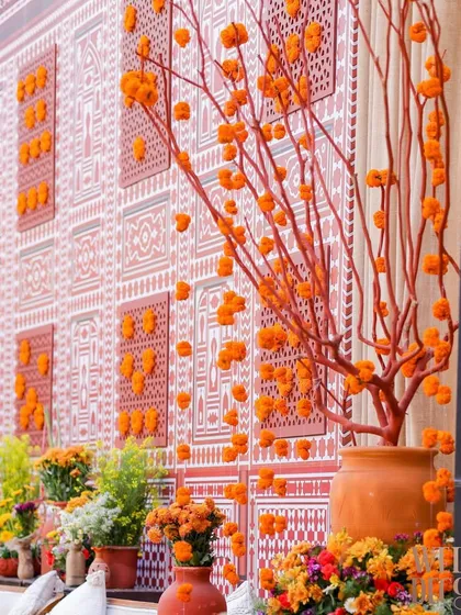A unique floral installation using dried branches painted terracotta and dotted with marigold flowers, set against a patterned backdrop. This added a modern artistic touch to the traditional theme.