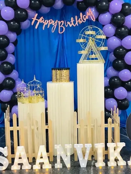 A black and purple balloon arch against a blue curtain, with white pedestals and marquee letters.