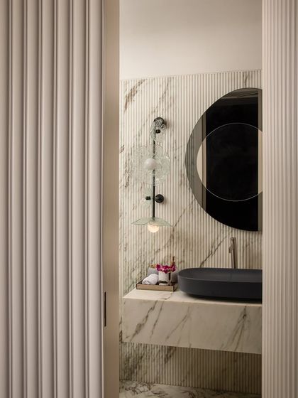 Inspired by Parisian style, this powder room exudes luxury and refinement. A peek through the fluted doorway reveals a space where rich marble textures, a dark stone basin, and warm ambient lighting create a lavish and elegant ambiance.