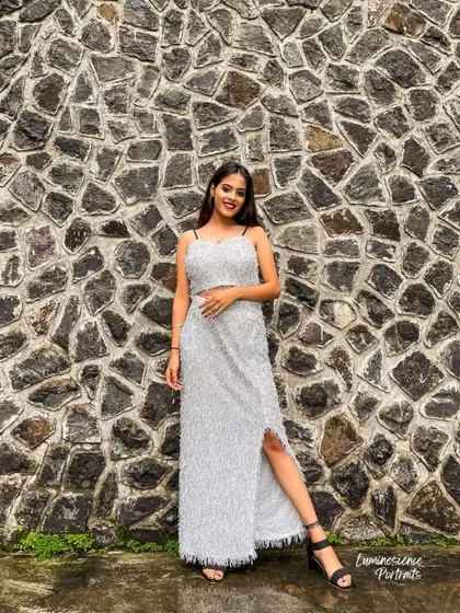 A full-length shot of the model smiling in her silver tinsel dress, set against a dark, textured stone wall.