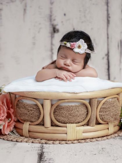 A baby girl with a floral headband resting in a beautiful wicker basket, surrounded by bouquets of pink and yellow flowers.