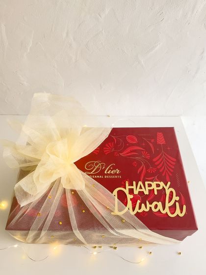 Our signature red Diwali gift box, elegantly wrapped in tulle with a golden "Happy Diwali" tag, illuminated by fairy lights.