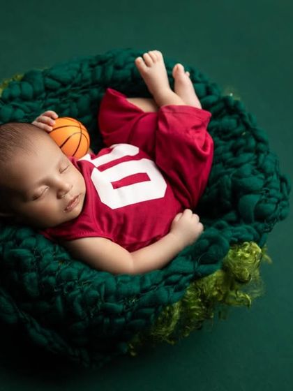 Our little basketball star, peacefully dreaming of future games. The baby is dressed in a red jersey and nestled in a green knit prop, holding a miniature basketball.