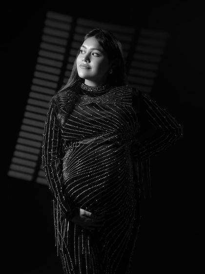 The same artistic lighting style, presented in black and white. This version emphasizes the texture of the gown and the thoughtful expression of the mom-to-be.